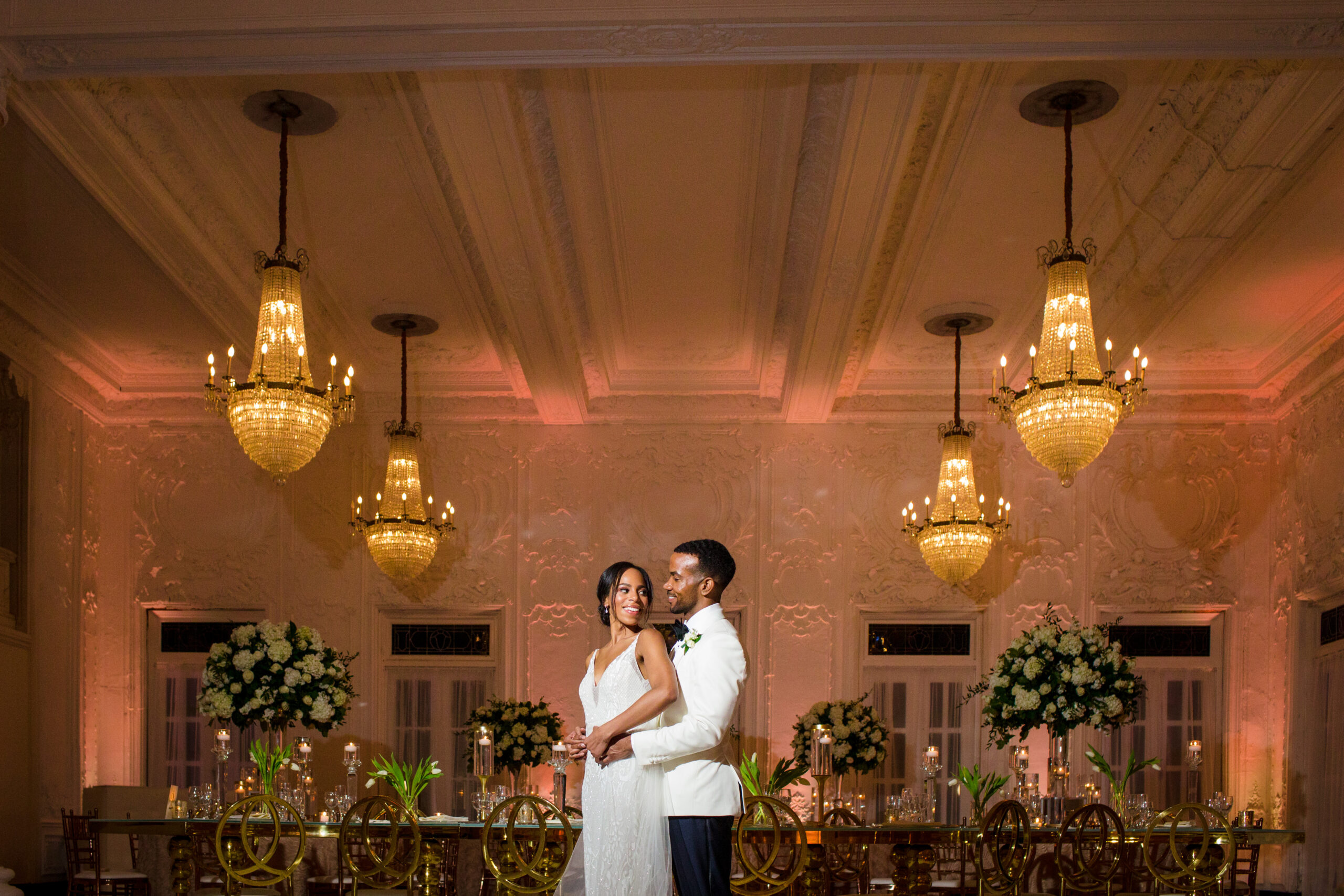 A bride and groom posing in front of chandeliers in a ballroom.
